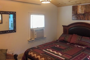 A bedroom with a bed, a window with an air conditioning unit, a mirror, and a wall-mounted artwork.