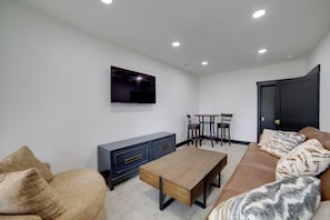A modern living room with a flat-screen TV mounted on the wall, a wooden coffee table, a sofa with patterned cushions, a dark-colored cabinet, and a dining table with chairs.