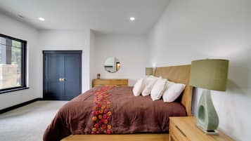 A bedroom with a large bed, a wooden headboard, a bedside table with a lamp, a dresser, a mirror, and a dark door.