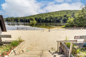 Ein Sandstrand mit einer Holzumzäunung, einem Schild für eine Zone sauberer Luft und einem See mit einem bewaldeten Hügel im Hintergrund.