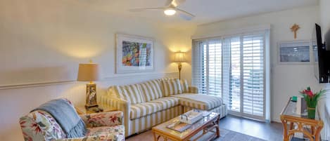 Una sala de estar con un sofá a rayas, un sillón con estampado floral, una mesa de centro, un ventilador de techo y un ventanal corredero.