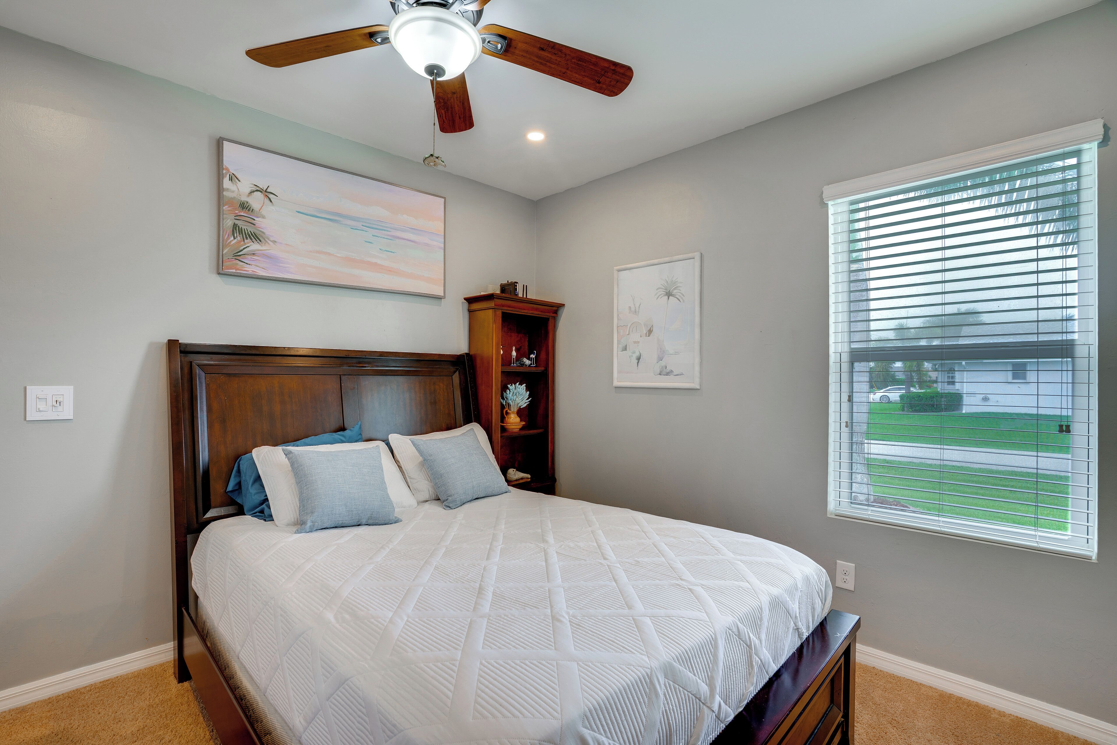 A bedroom with a bed, a ceiling fan, a window with blinds, and artwork on the walls.
