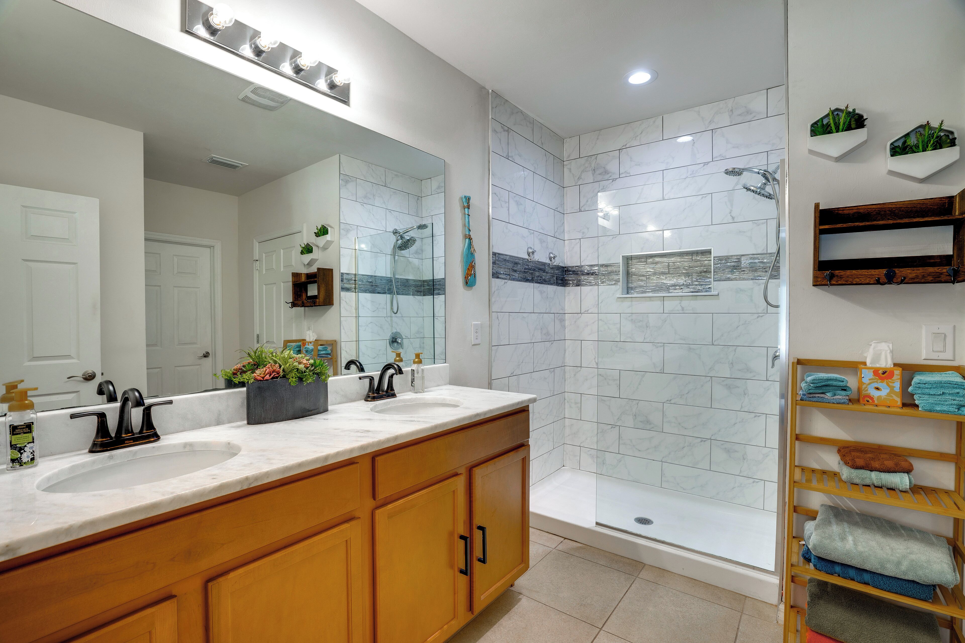 Une salle de bain moderne avec un grand miroir, deux vasques, un espace douche et un porte-serviettes avec des serviettes pliées.