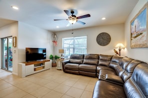 A modern living room with a leather sofa, a flat-screen TV, a ceiling fan, and a large wall clock. - Pet-friendly Cape Coral Home w/ Pool & Hot Tub (Cape Coral)