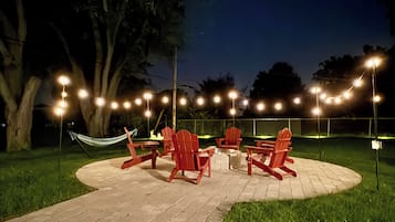 Un área de estar al aire libre con sillas rojas y una mesa, rodeada por un camino de ladrillo y luces colgantes.