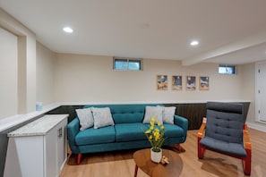 A living room with a teal sofa, a wooden coffee table, and a grey armchair. There are four framed artworks on the wall and a window above the sofa. - Walk to Beach! Mccullom Lake Family Getaway (McHenry)