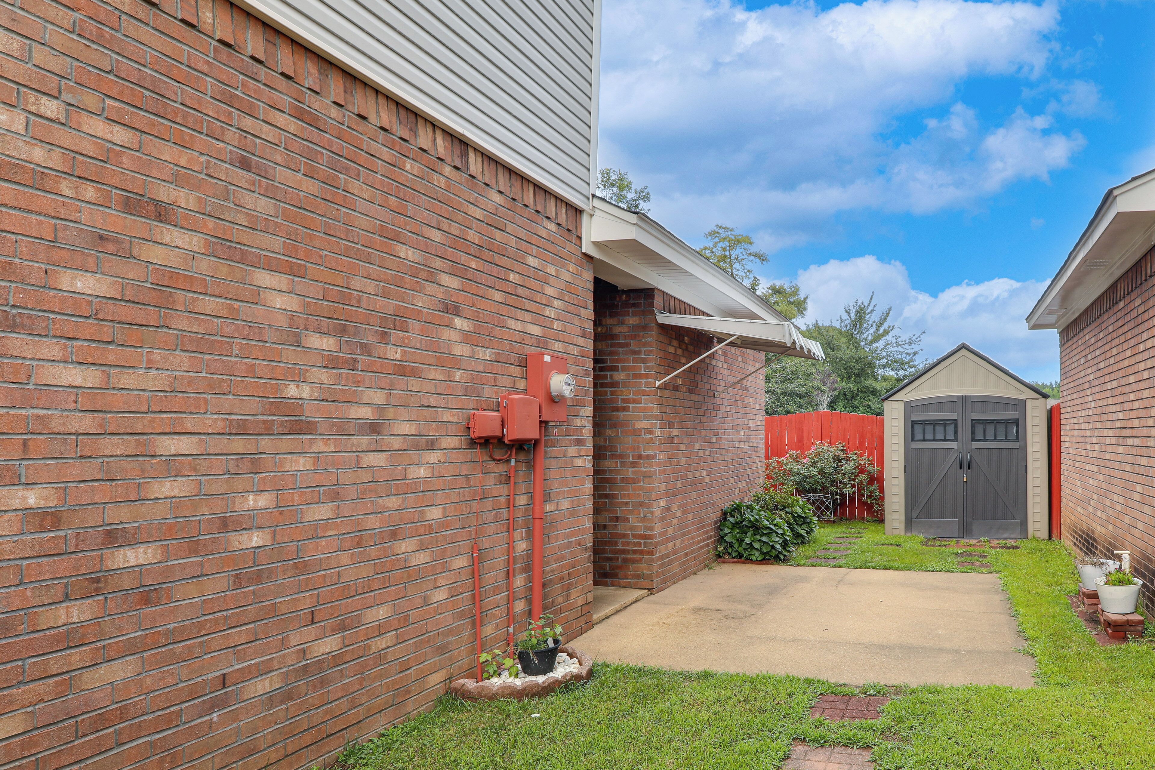 A brick building with a covered porch, a small garden with a potted plant, and a shed with a dark door.