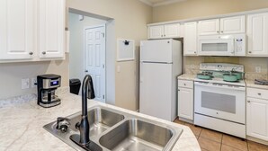 Una cocina con armarios blancos, un lavamanos de acero inoxidable, una cafetera, un refrigerador, un microondas y un horno.