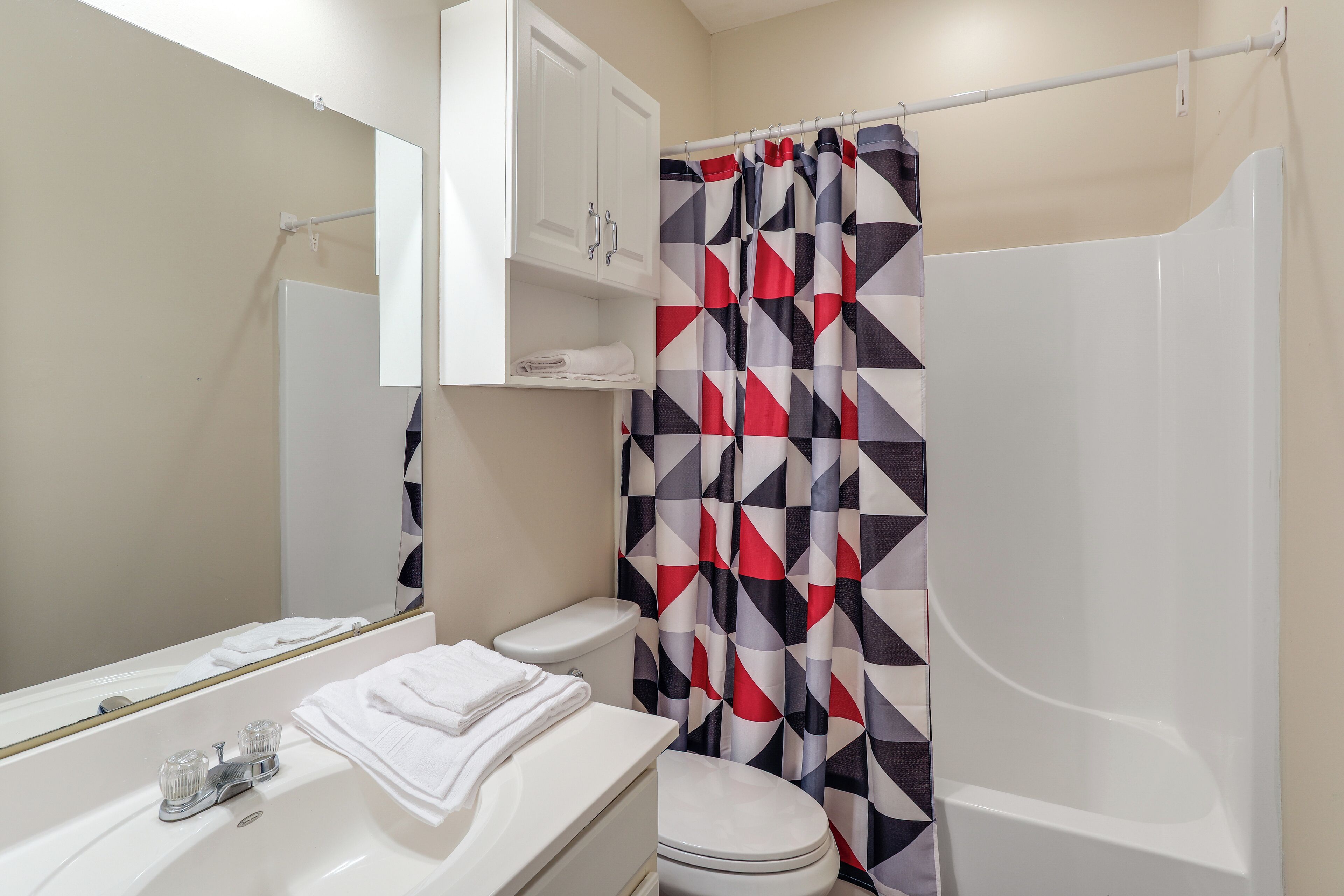 A bathroom with a white toilet, a sink with a glass soap dispenser, a mirror, and a shower curtain with a geometric pattern.