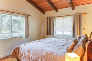 Um quarto com uma cama grande, duas janelas com cortinas, teto de madeira e uma luminária de cabeceira.