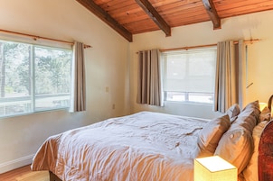 Un dormitorio con una cama grande, dos ventanas con cortinas, techo de madera y una lámpara de noche.
