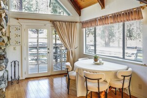 Una habitación con pared de piedra, puerta de madera, ventana con persianas y una mesa redonda con sillas, con vista a árboles fuera.