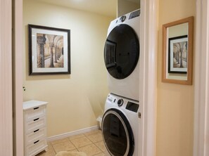 A laundry room with two stacked washing machines, a white cabinet with drawers, and two framed pictures on the wall. - Snowbird Desert Condo - Tennis Garden, Golf, 2BR/2BA (La Quinta)