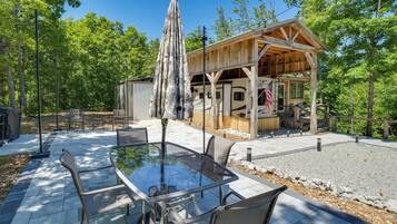 A covered outdoor seating area with a table and chairs, a wooden structure with a roof, and a recreational vehicle in the background.