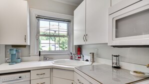 Una cocina con armarios blancos, una ventana, cafetera, un lavaplatos y microondas.