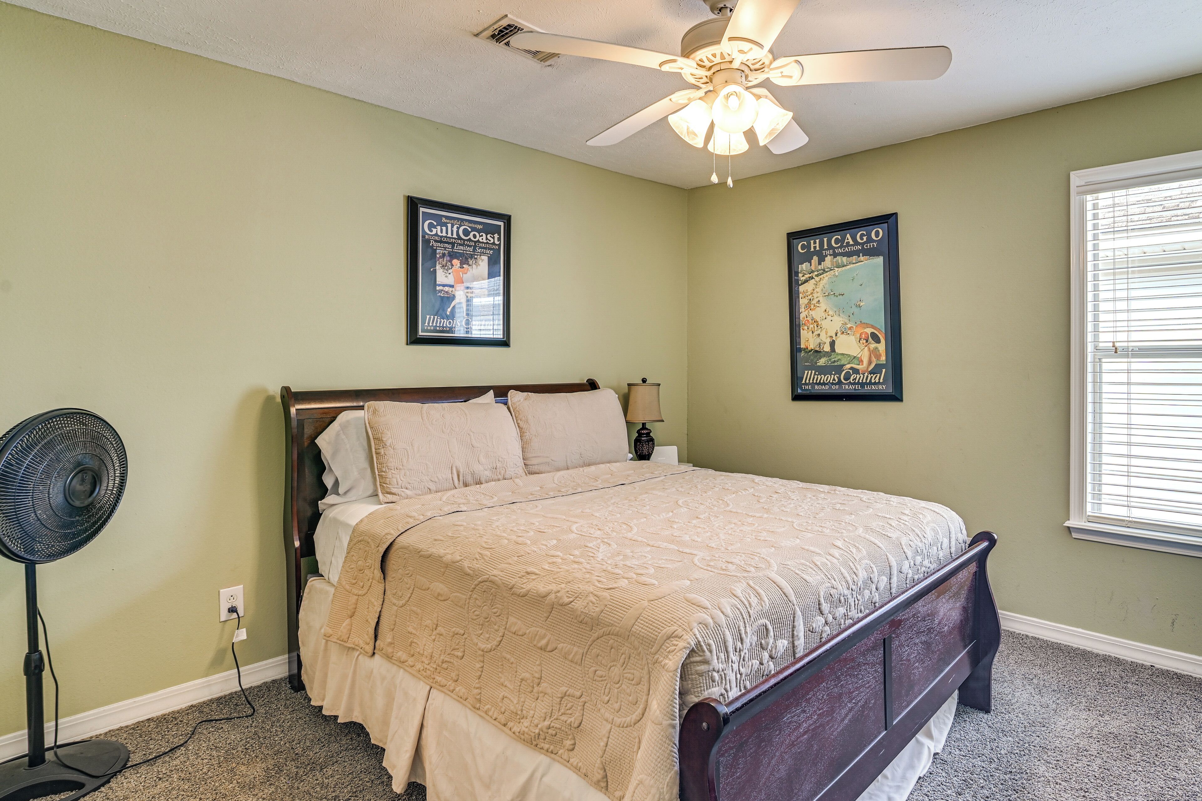 A bedroom with a bed, a ceiling fan, a standing fan, and two travel posters on the wall.