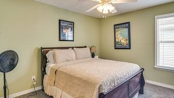A bedroom with a bed, a ceiling fan, a standing fan, and two travel posters on the wall.
