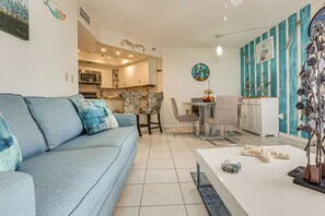 Ein modernes Wohnzimmer mit einem blauen Sofa, einem Essbereich mit einem Glastisch und einer Küche im Hintergrund.