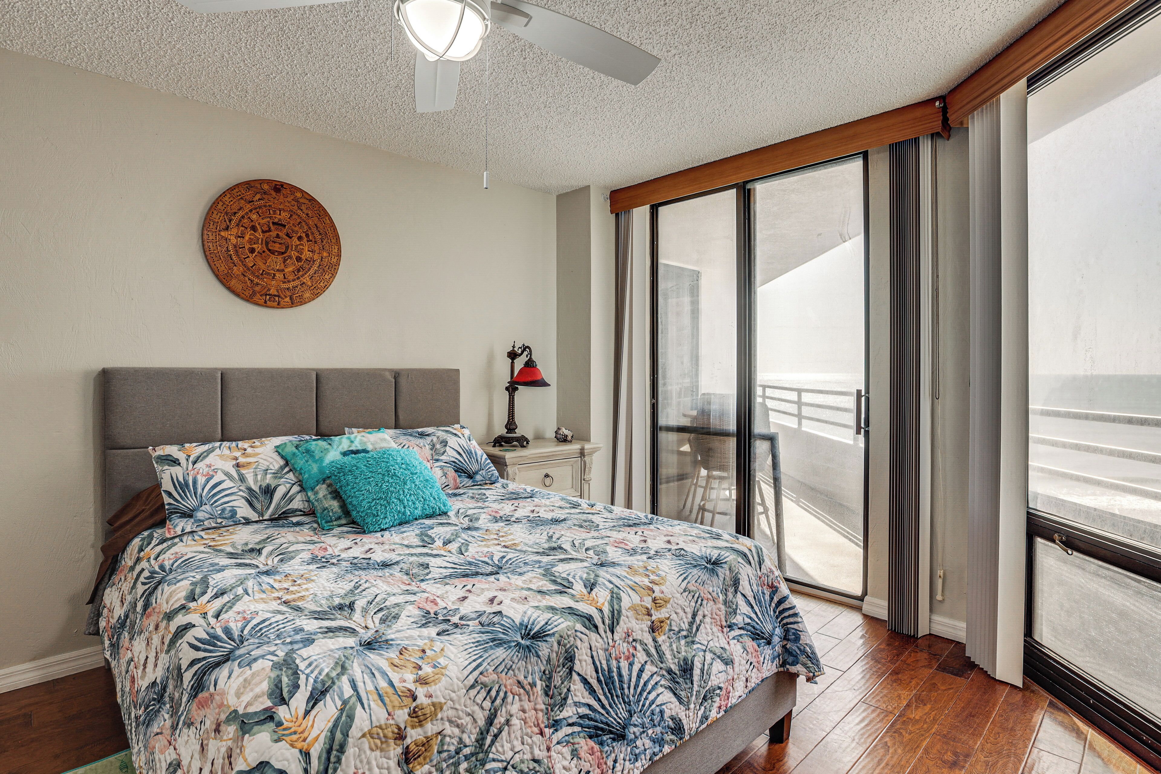 Une chambre avec un grand lit, un ventilateur de plafond, un élément de décoration circulaire au mur, une table de chevet et une vue à travers une porte-fenêtre coulissante.