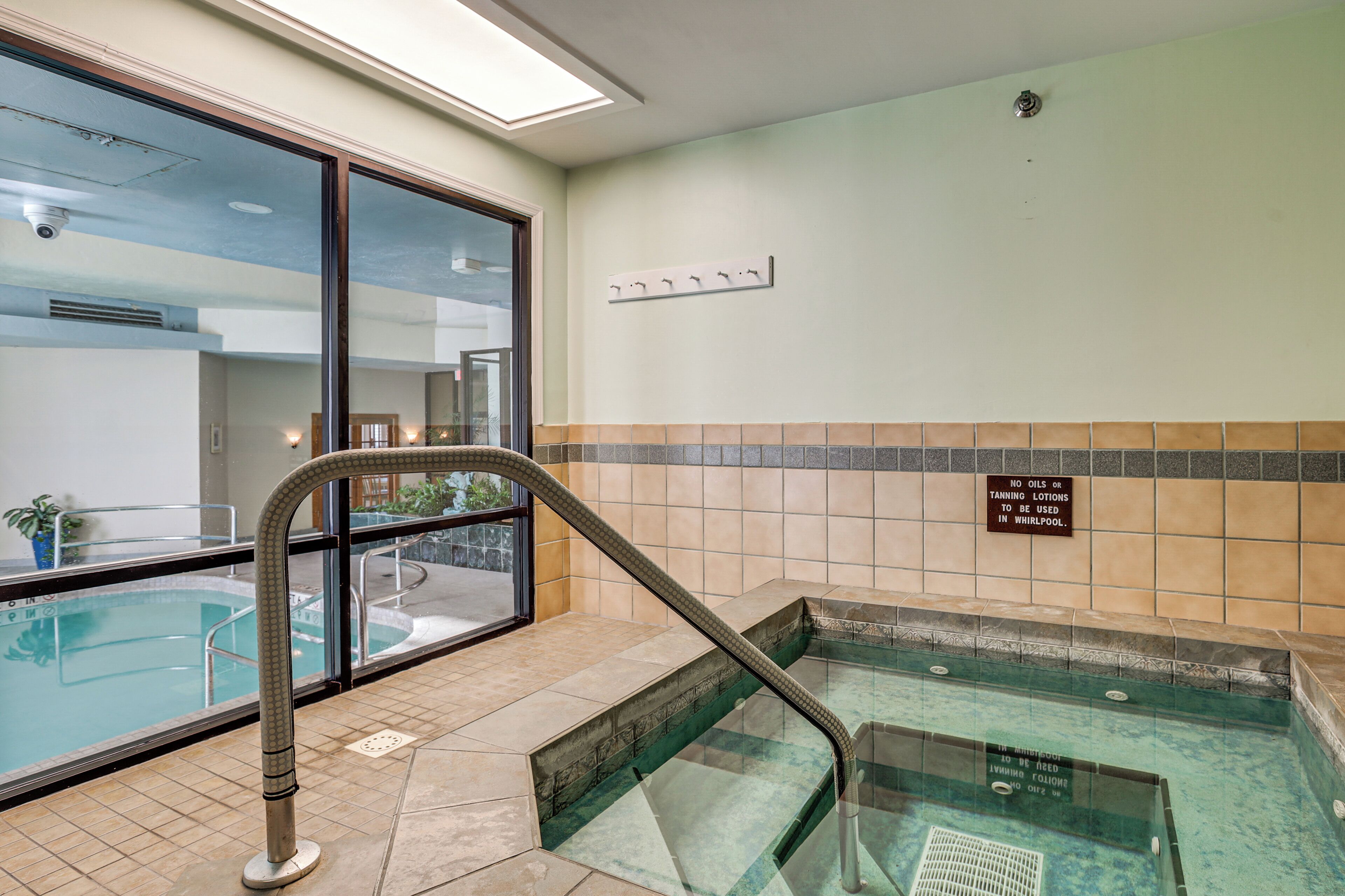 A pool area with a glass barrier, a sign prohibiting oils or tanning lotions in the whirlpool, and a tiled wall with a warning sign.