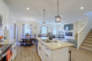 A modern kitchen with a central island, a dining area with a wooden table and chairs, a living room with a sofa and a TV, and a staircase leading to an upper level. - Updated Family Home w/ Patio in Brighton (Brighton)