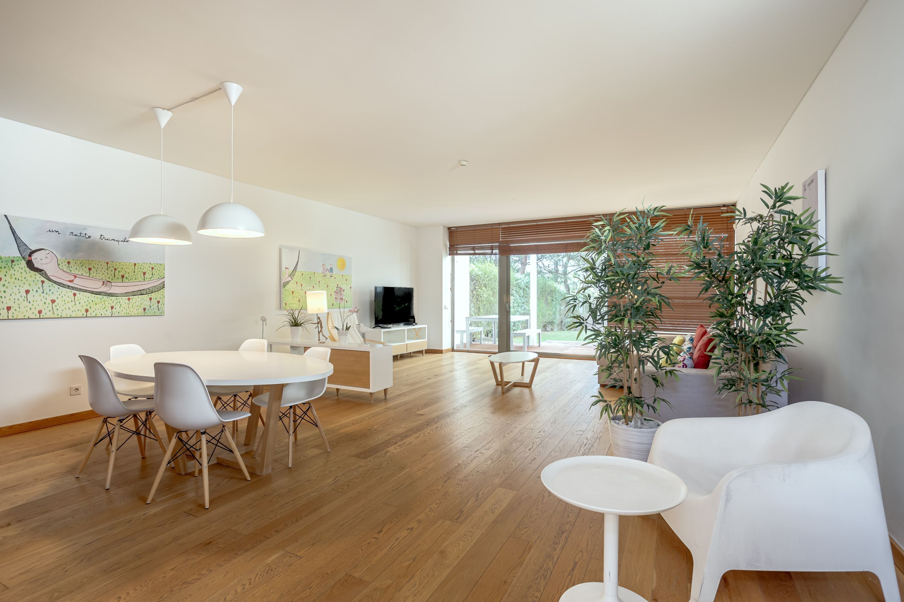 Una sala de estar moderna con comedor, suelo de madera, muebles blancos y un ventanal con persianas.
