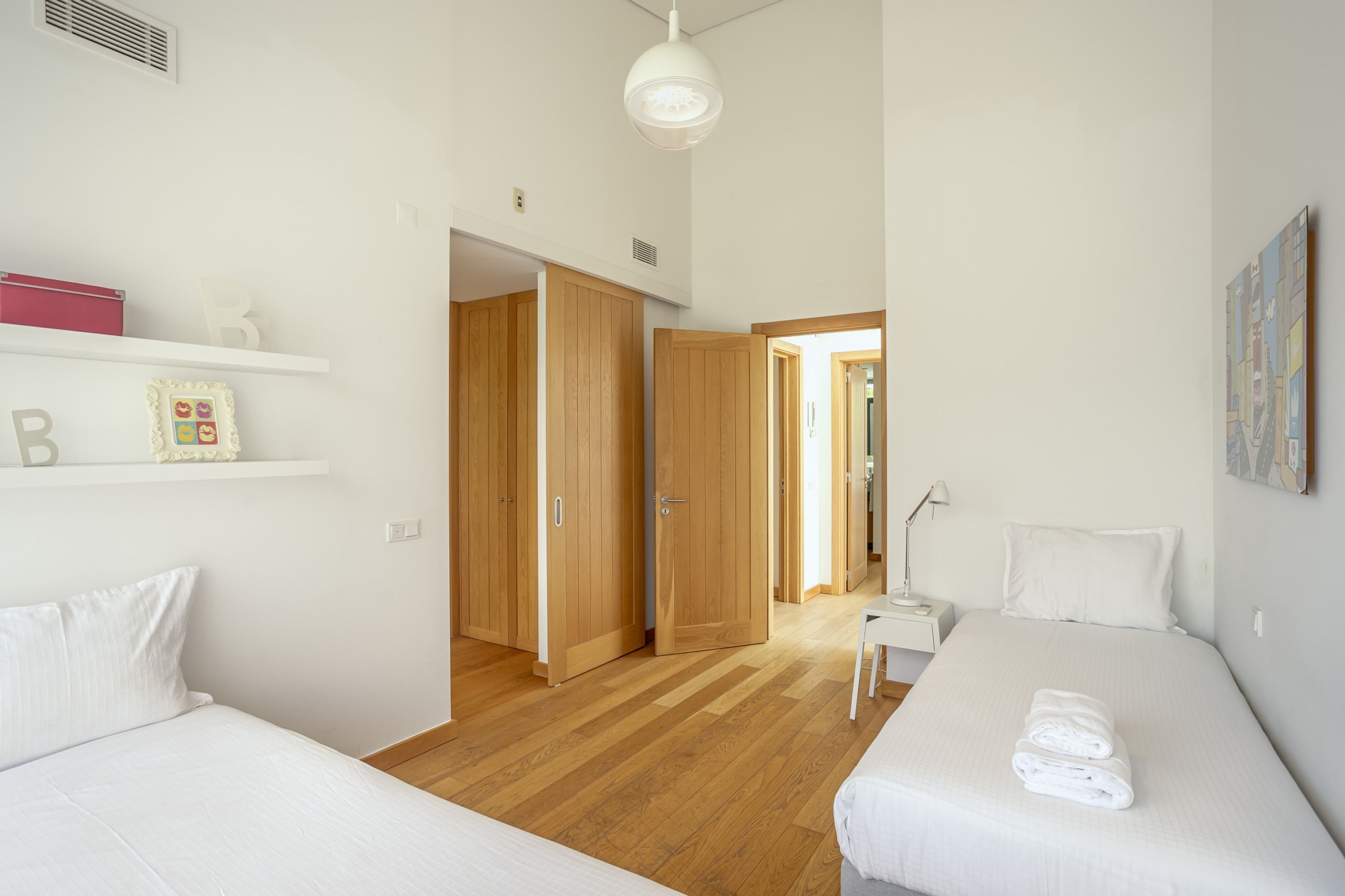 Quarto moderno com duas camas, piso de madeira, parede branca e um corredor que dá para outro quarto.