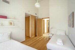 Quarto moderno com duas camas, piso de madeira, parede branca e um corredor que dá para outro quarto.