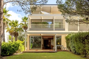 A modern two-story house with a large glass door entrance, a balcony, and a well-maintained garden. - Troia Atlantic Villa by Homing (Grandola)