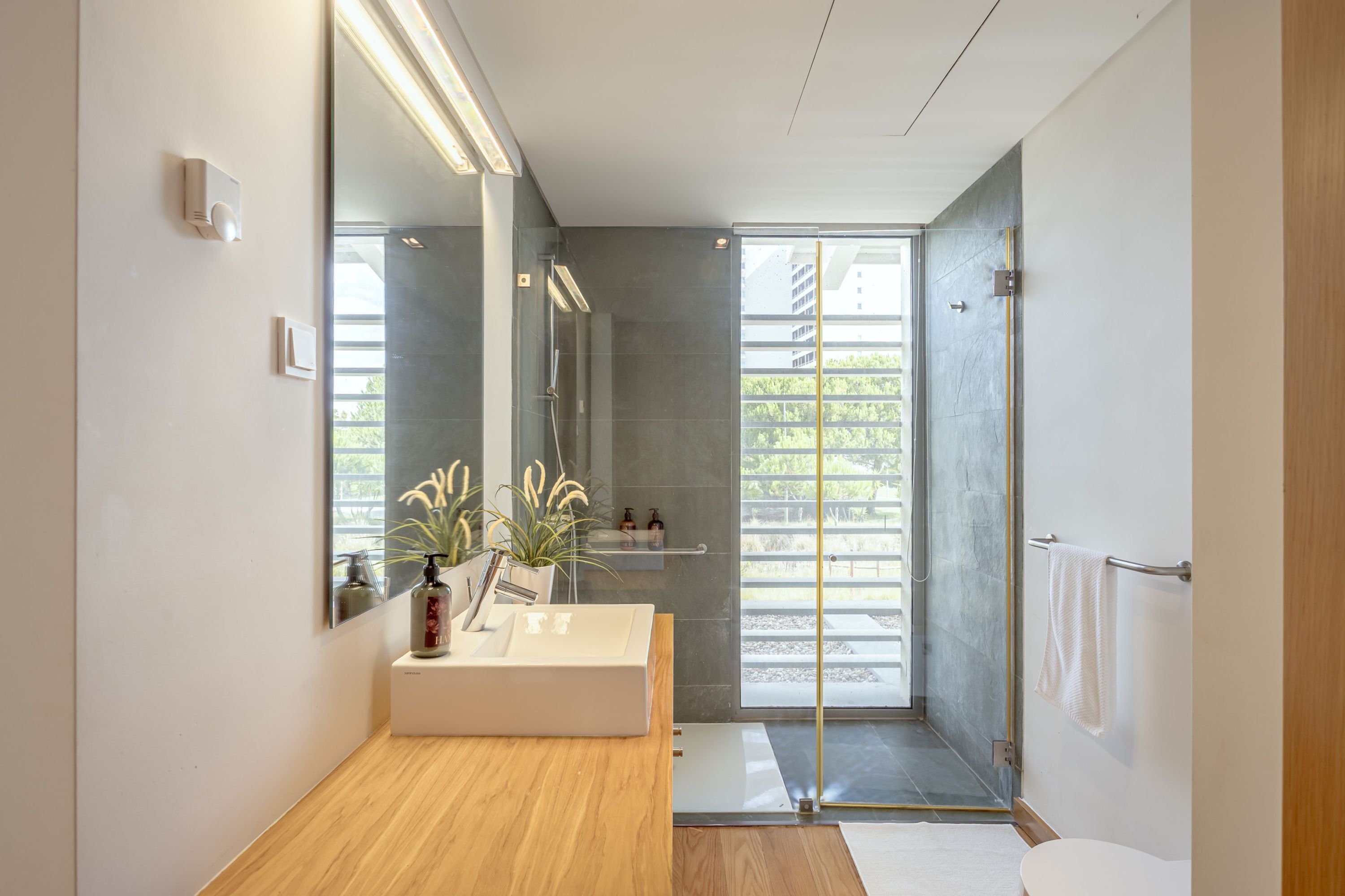 A modern bathroom with a large mirror, a wooden vanity, a shower with a glass door, and a view of greenery outside.