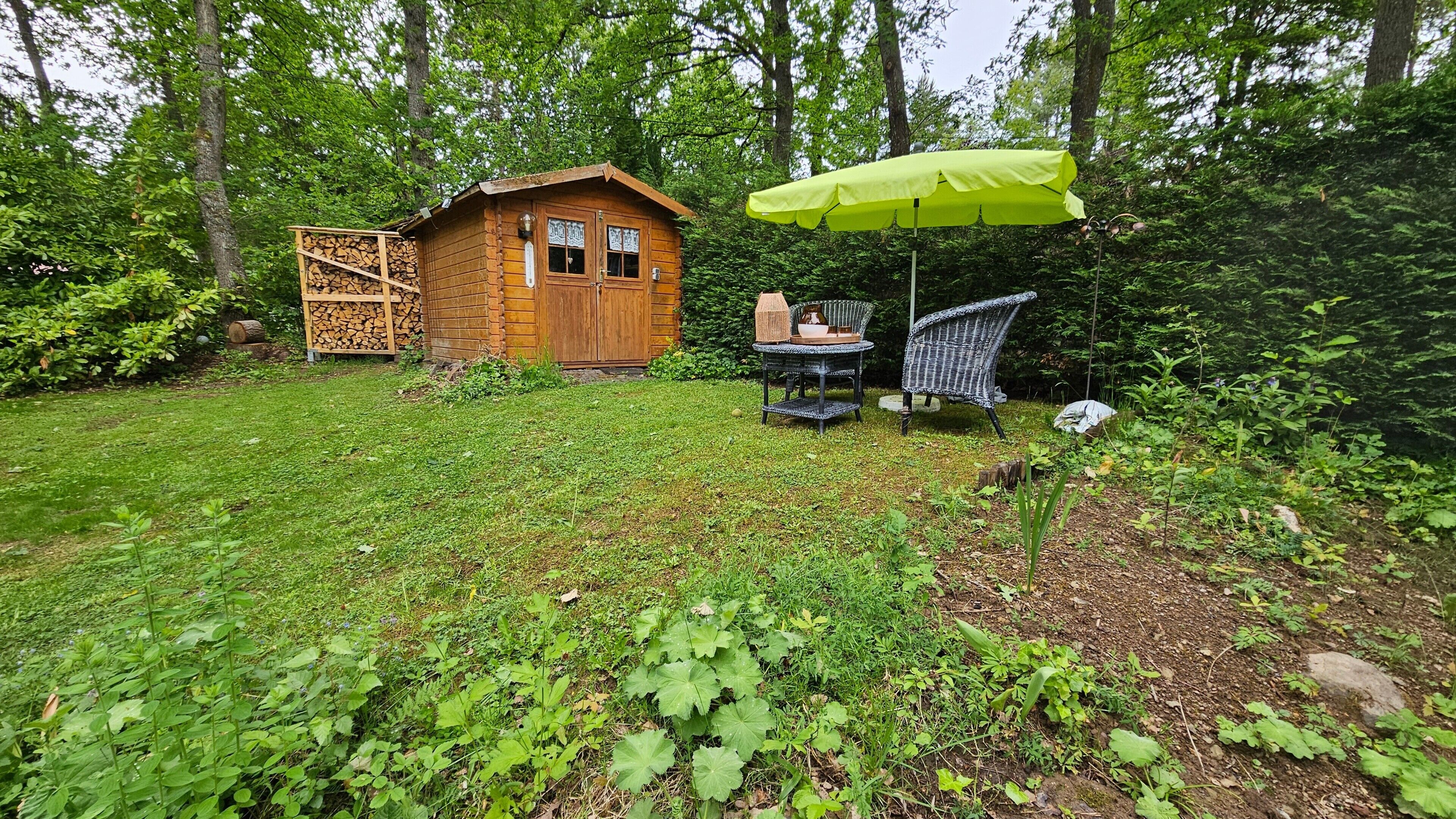 Eine Holzhütte mit einem grünen Sonnenschirm, einem Korbstuhl und einem Tisch mit einer Vase und einer Schüssel.