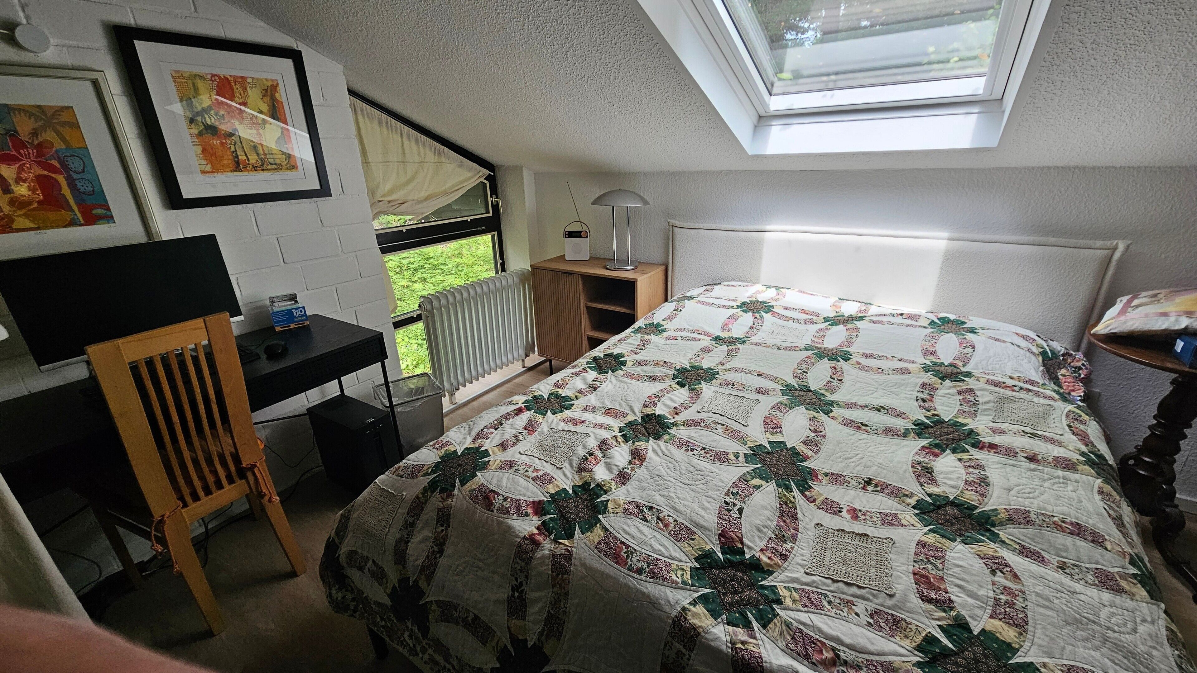 Ein Schlafzimmer mit einem Bett, einem Schreibtisch, einem Stuhl, einem Heizkörper und einem Fenster mit Blick auf Grünflächen.