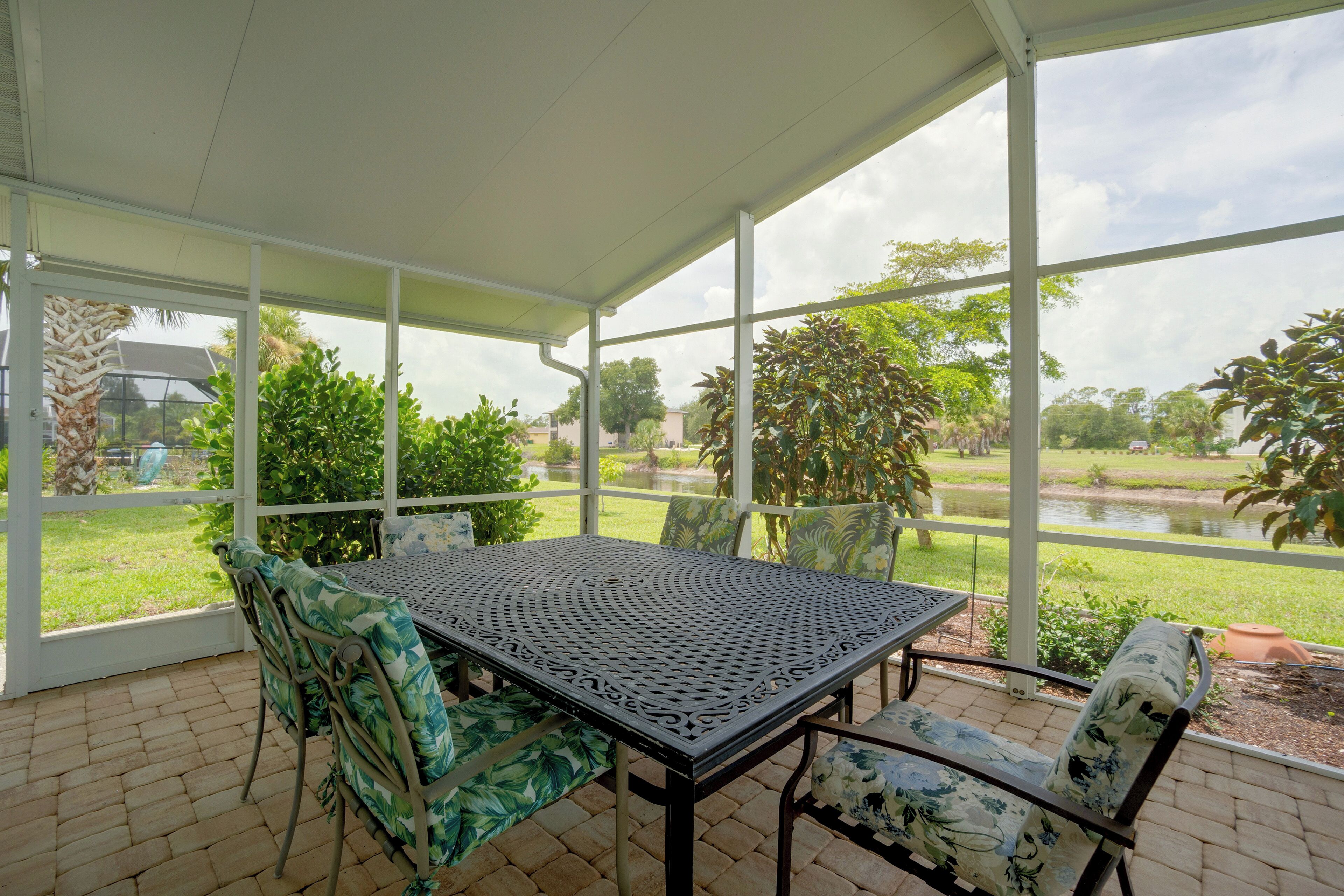 Un patio cubierto con mesa y sillas, ventanas grandes y vistas a un estanque y vegetación.