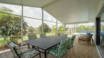 A covered patio with a table and chairs, a pool, and a view of a garden and water body.
