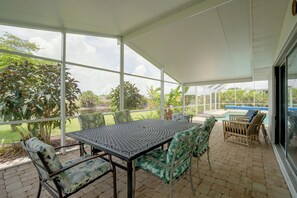 A covered patio with a table and chairs, a pool, and a view of a garden and water body.