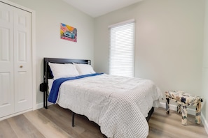 Quarto com uma cama, uma porta branca, uma janela com persianas e um pequeno banco.