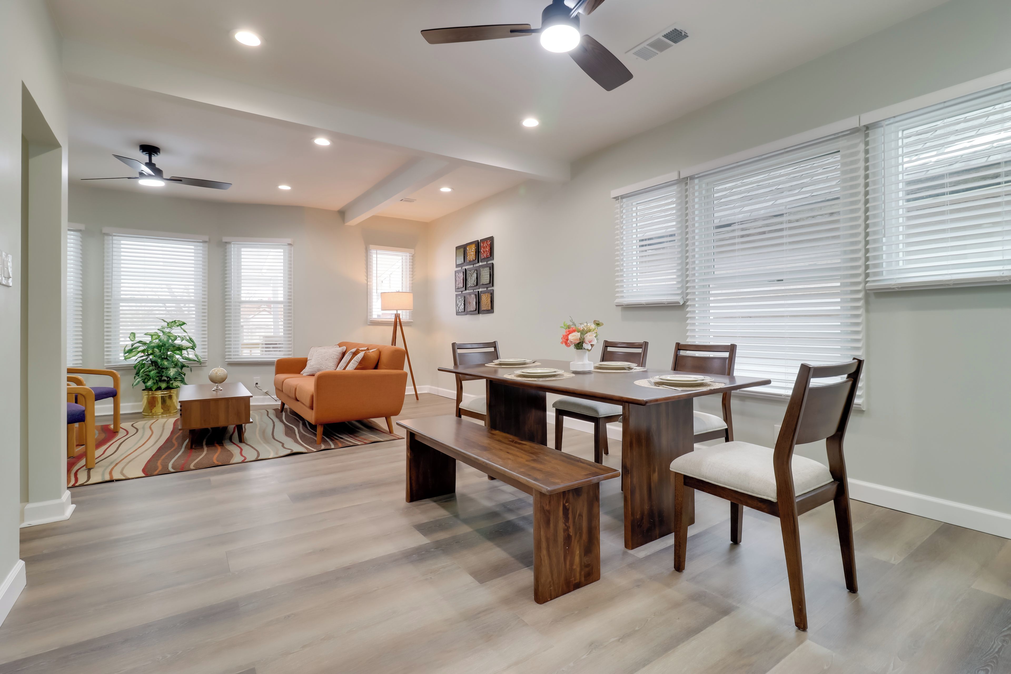 A spacious living area with a dining table, benches, and a sofa. There are ceiling fans, recessed lighting, and large windows with blinds.