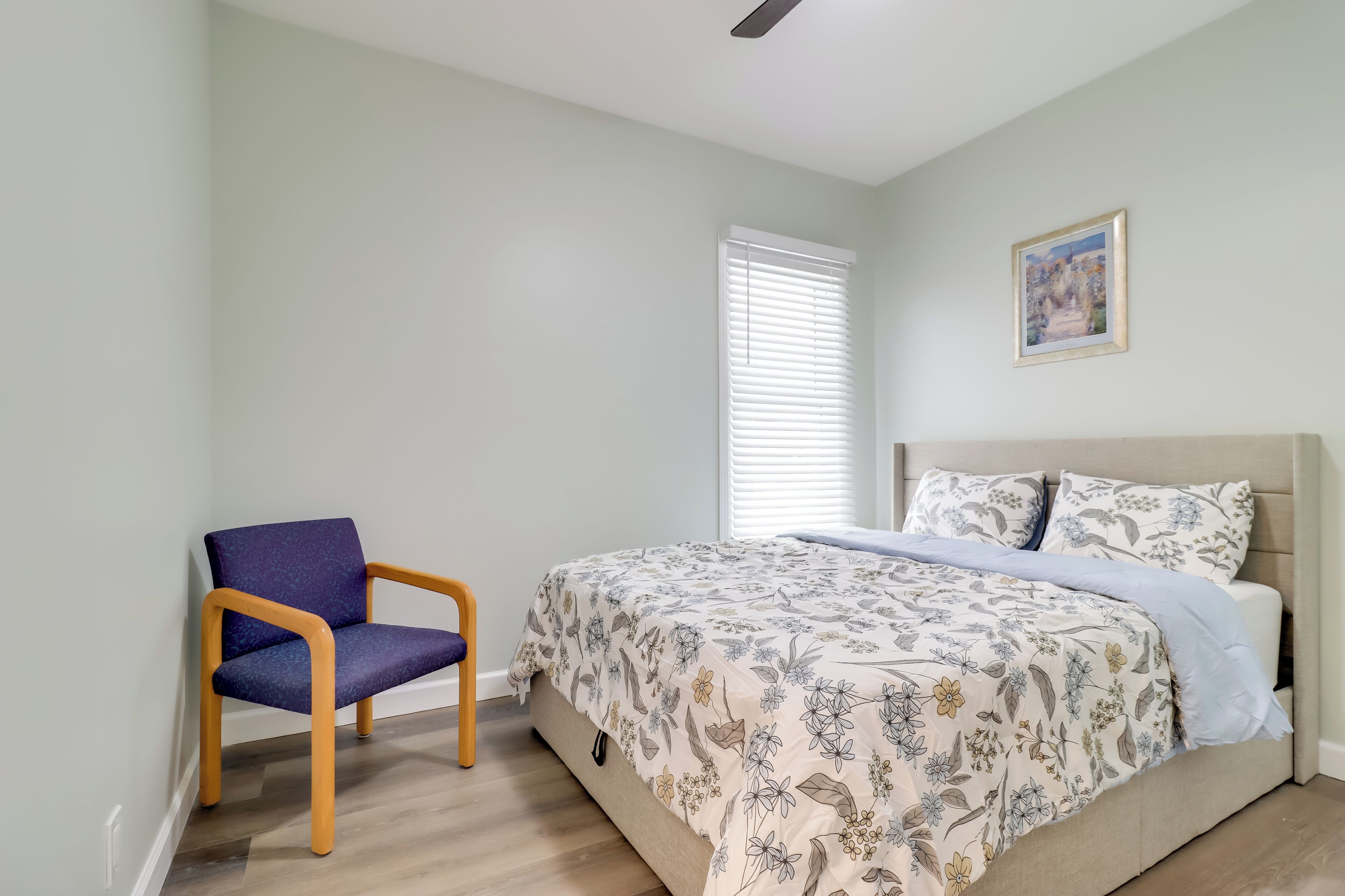 A bedroom with a bed, a chair, a window with blinds, and a painting on the wall.