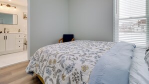 Ein Schlafzimmer mit einem Bett, einem Sessel, einem Frisiertisch mit Spiegel und einem Fenster mit Jalousien.