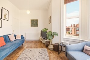 A living room with a blue sofa, a TV, a plant, and a window with a view of houses. - Sunset Views - North East Escapes (Tynemouth)