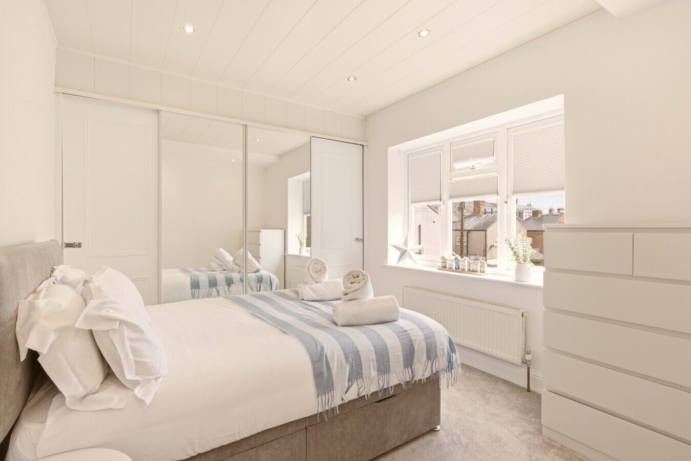 A bedroom with a bed, a dresser, a mirror, and a window with a view of buildings.
