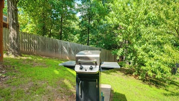 Una parrilla para barbacoa con panel de control y superficie de cocción plana, ubicada en un patio césped con valla de madera y árboles al fondo.