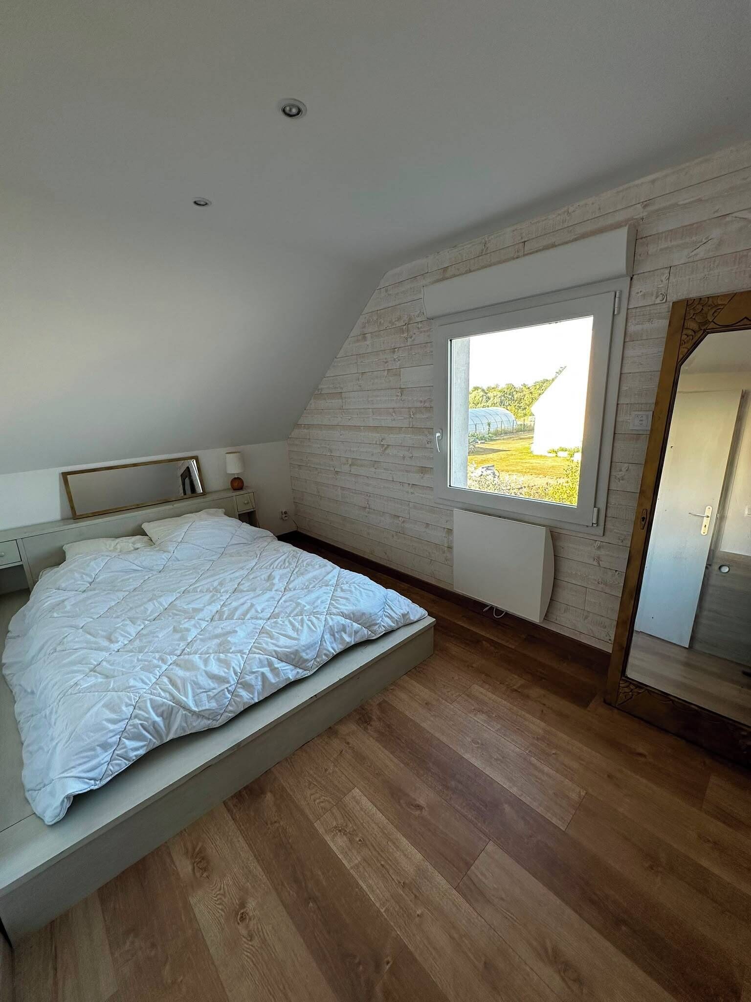Ein Schlafzimmer mit schrägen Wänden, einem Bett mit weißer Decke, einem Fenster mit Blick auf Grünflächen und Holzboden.