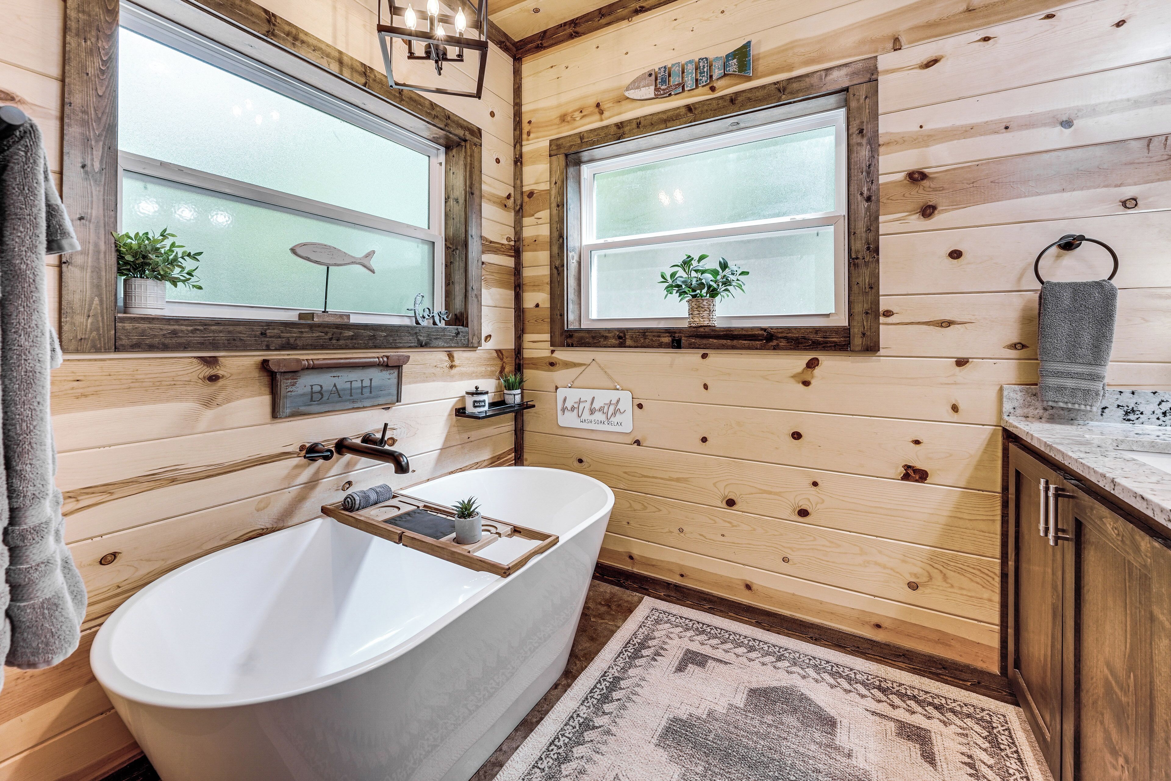 A wooden bathroom with a freestanding bathtub, two windows with potted plants, and a towel rack.