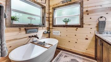 A wooden bathroom with a freestanding bathtub, two windows with potted plants, and a towel rack.