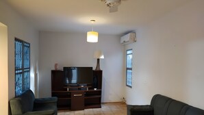 Una sala de estar con una televisión de pantalla plana, dos sofás, una mesa de centro, un ventilador de techo y una ventana con persianas.