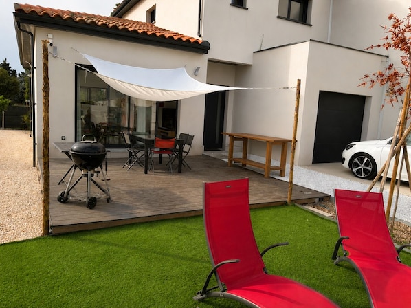 Eine Terrasse mit Grill, Gartenmöbeln und einem weißen Auto, das in einer Wohngegend geparkt ist.