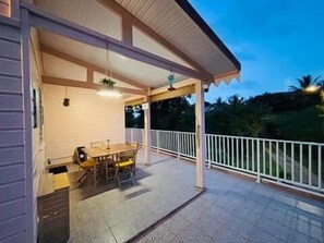 Un espacio al aire libre techado con una mesa y sillas, baranda blanca y vistas de la vegetación circundante.