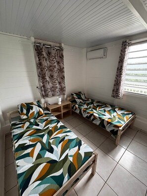 Una habitación con dos camas de madera con ropa de cama de patrones geométricos coloridos, una mesita de noche de madera, una ventana con cortinas estampadas y un techo blanco con líneas horizontales.