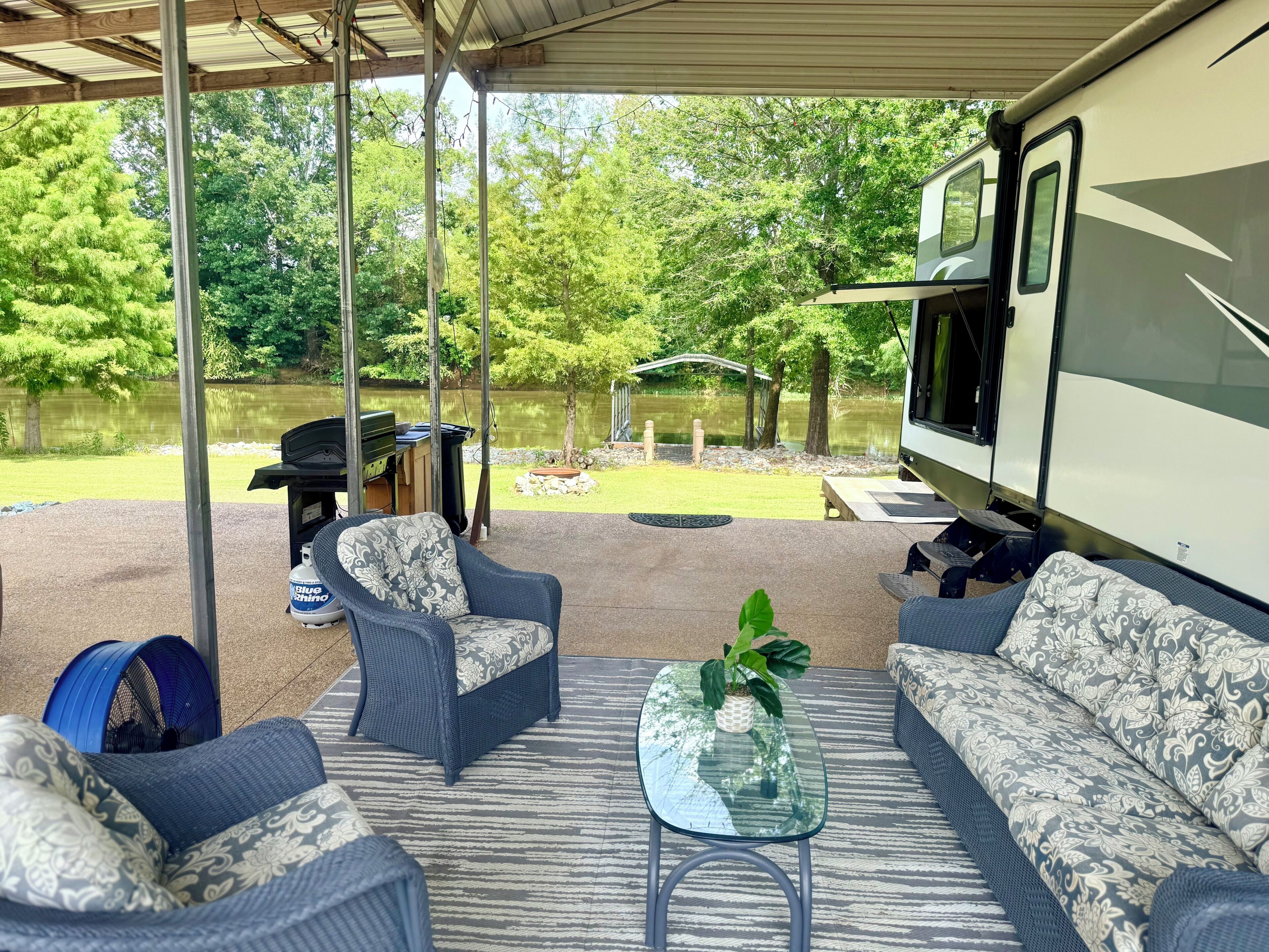 A covered outdoor area with a sofa, chairs, and a table, overlooking a lake and trees.
